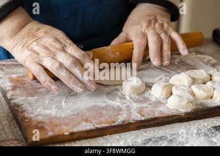 Le mani di un panettiere anziano preparano cerchi di impasto usando un rullo su una superficie polverosa, evidenziando un processo di cottura rustico e l'arte della creazione tradizionale Foto Stock