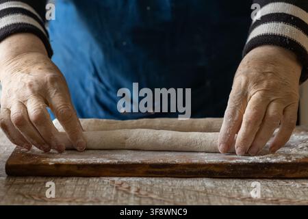 Mani anziane arrotolano pasta fresca su una superficie infarinata per dimostrare l'abilità del pane classico, evidenziando tecniche ereditate e il calore di au Foto Stock