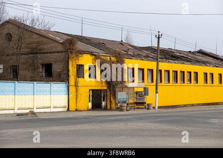 Horodok, Ucraina – 23 marzo 2026: Una struttura industriale abbandonata con il tetto crollato e vite cresciute spontaneamente si trova accanto a una fermata dell'autobus, illustrando l'ambi Foto Stock