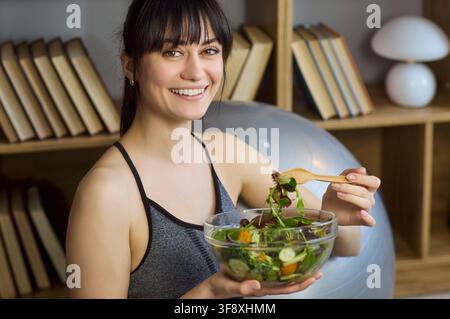 Giovane donna felice con una ciotola di insalata a casa, alimentazione sana e stile di vita Foto Stock