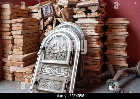 Porte di stufa in ghisa antiche ornate appoggiate a pile di mattoni e tegole di argilla patinate all'interno di una bottega o magazzino rustico. Il compagno invecchiato Foto Stock