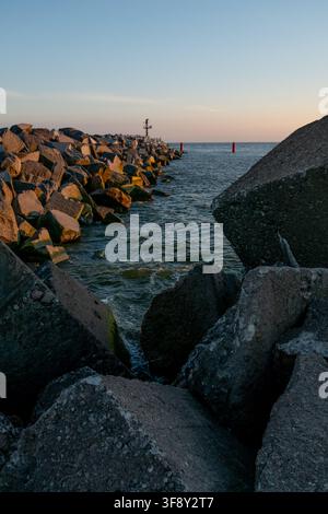 Grandi barriere di mare in cemento e rocce lungo una costa al tramonto, con acqua calma e una boa segnaletica rossa in lontananza. Foto Stock
