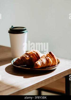 Due croissant dorati e friabili serviti su un piatto di ceramica con una tazza di caffè da asporto, illuminati da una calda luce naturale su un tavolo di legno Foto Stock