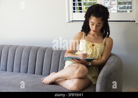 Donna seduta su divano grigio vicino alla finestra, indossa un vestito giallo, scrive in un taccuino blu verdastro con una matita Foto Stock