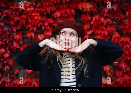 Donna alla moda in caldo abbigliamento autunnale che posa davanti a foglie rosse brillanti, concetto di stile di vita stagionale. Foto Stock
