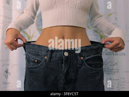 Donna in piedi che tira su la cintura dei suoi vecchi jeans oversize, mostrando il progresso significativo e il successo del suo percorso di perdita di peso e celebrando Foto Stock