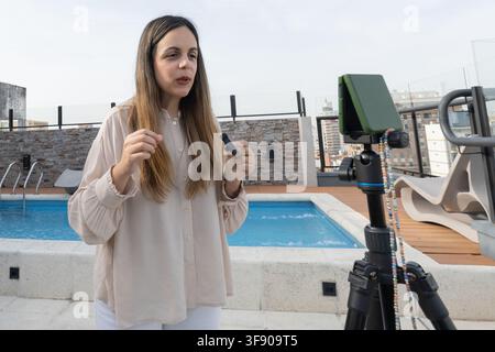 Prima piano di una donna che tiene un piccolo microfono lavalier mentre parla alla telecamera, smartphone montato su treppiede, piscina e skyline della città alle sue spalle Foto Stock