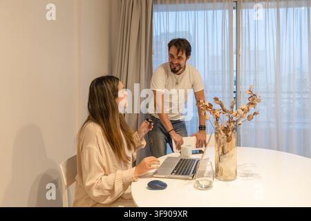 Donna seduta al tavolo che tiene un piccolo microfono lavalier mentre un uomo sorridente le sta accanto vicino alla finestra, laptop ed eucalipto essiccato sul tavolo Foto Stock