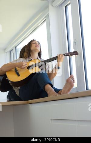 Donna africana seduta su una panca di legno sul davanzale di una finestra in un appartamento moderno e luminoso che suona la chitarra acustica Foto Stock
