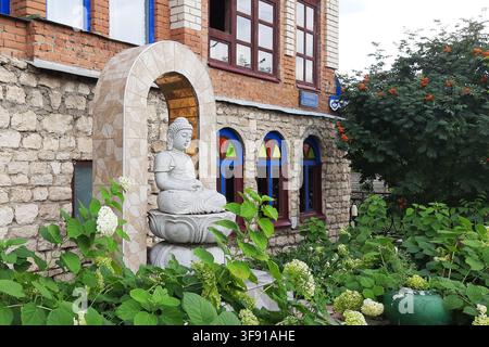 Sviyazhsk, Russia – 24 luglio 2026: Statua di Buddha tra le ortensie accanto a un edificio in mattoni ornato Foto Stock