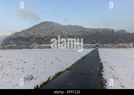 Strada di campagna attraverso campi coperti di neve e un villaggio rurale giapponese in inverno Foto Stock