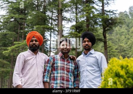 Scatto a bassa angolazione, dal busto in su, di tre amici maschi, tra cui due uomini sikh, in piedi insieme, che guardano l'obiettivo e sorridono mentre posano. Foto Stock