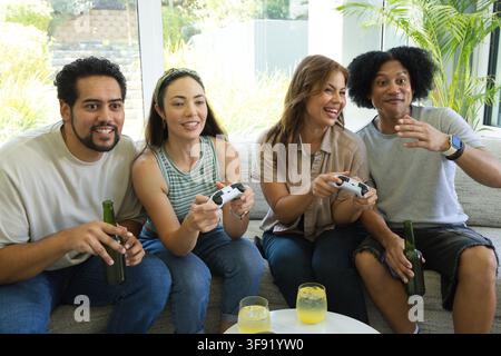 Amici diversi seduti su un divano grigio in salotto che giocano con controller bianchi, tenendo birra Foto Stock