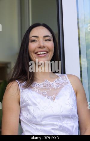 Donna sorridente, in piedi sulla porta, indossa top di pizzo bianco a fiori, porta in vetro che riflette il cielo Foto Stock