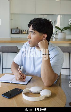 Uomo che scrive a un tavolo di legno in cucina tenendo una penna sopra un taccuino, smartphone, cuffie, con maglietta blu Foto Stock