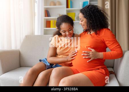 Madre incinta nera sorridente e figlia che si legano insieme, la ragazza tocca la pancia della mamma Foto Stock