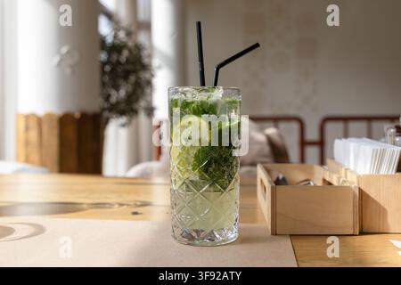 Rinfrescante bevanda mojito piena di ghiaccio, menta e lime si posa su un tavolo di legno caldo in un caffè illuminato dal sole, evocando un'atmosfera estiva fresca per l'ospitalità Foto Stock
