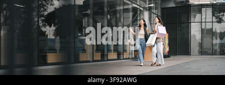 Due donne asiatiche che camminano fuori dal centro commerciale con borse di carta per le offerte del Black Friday Foto Stock