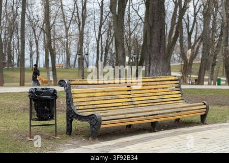 Una panca da esterno invecchiata si trova accanto a un contenitore per i rifiuti su un sentiero erboso tranquillo all'interno di un bosco pubblico, simboleggiando relax e ghirlanda comunitaria Foto Stock