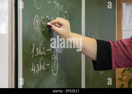 Un educatore tiene la gesso mentre calcola equazioni matematiche su una lavagna per coinvolgere i bambini che imparano in un ambiente di classe primaria focalizzato Foto Stock