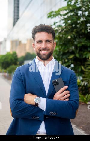Ritratto di un uomo d'affari sorridente che indossa un blazer blu, tiene uno smartphone e sta con le braccia incrociate davanti a un edificio moderno di uffici. Foto Stock