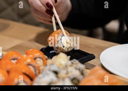 Primo piano di una mano curata che usa le bacchette per tenere un rotolo di salmone sopra una ciotola di salsa in un ristorante, catturando il fascino sensoriale e il culto culinario Foto Stock