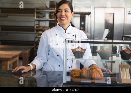 Pasticcera che mostra croissant e muffin in stampi a tulipano alla vetrina del forno, indossando una giacca da chef Foto Stock