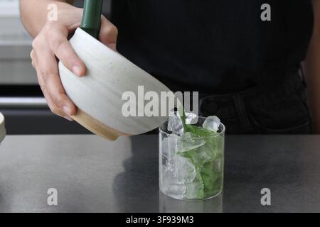 Una persona versa con cura il tè verde matcha da una chawan di ceramica in un bicchiere trasparente contenente cubetti di ghiaccio per preparare un latte. Foto Stock