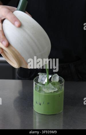 Una persona versa con cura il tè verde matcha da una chawan di ceramica in un bicchiere trasparente contenente cubetti di ghiaccio per preparare un latte. Foto Stock