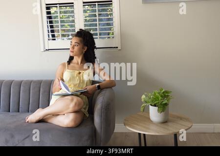 Donna adulta seduta su un divano grigio trapuntato vicino alla finestra, tiene un quaderno aperto e indossa uno smartwatch Foto Stock