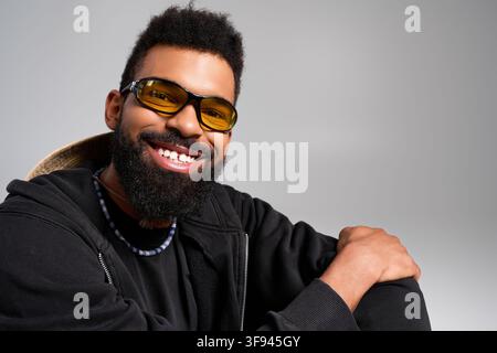 Un bel giovane con abiti comodi e occhiali da sole alla moda sorride calorosamente mentre è seduto. Foto Stock