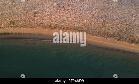 Vista aerea con drone di una linea di costa sabbiosa strutturata che incontra acque turchesi calme, con onde gentili che si infrangono lungo la costa. Foto Stock