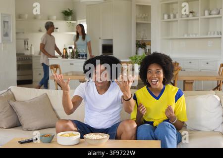 Donne afroamericane che tifano sul divano a casa in maglietta e maglia con popcorn, patatine e telecomando Foto Stock