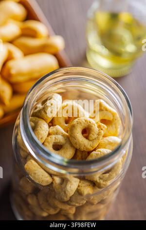 Snack croccanti a forma di anello (taralli) ammucchiati in un barattolo di vetro trasparente su un tavolo di legno, evidenziando la consistenza, il colore dorato e uno spuntino salato fatto in casa, perfetto per antipasti o per mangiare con tranquillità. Foto Stock