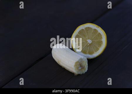 Banana sbucciata e mezza limone appoggiati su una superficie di legno scuro e rustico, rappresentanti opzioni di snack salutari Foto Stock