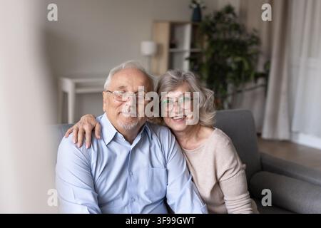 Coppia di anziani sorridenti che si abbracciano sul divano in soggiorno, ritratto Foto Stock