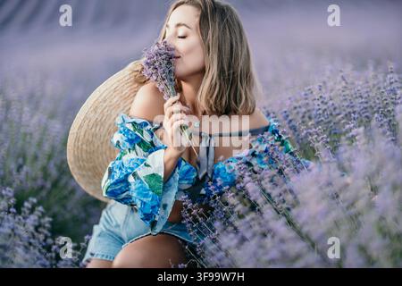 Donna campo di lavanda sorridente giovane donna che annusa un mazzo di fiori viola mentre è seduta in un campo estivo in fiore Foto Stock