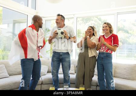 Amici diversi che esultano e applaudono in soggiorno indossando maglie, tenendo un pallone da calcio e una bandiera Foto Stock