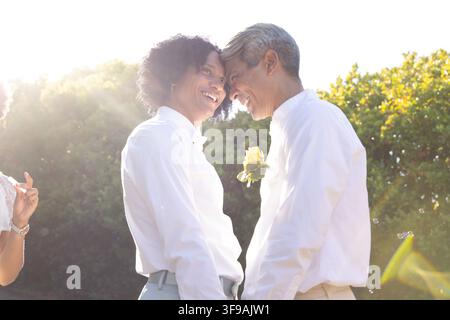 Coppia afroamericana che si tocca la fronte, sorridente in giardino, indossa camicie bianche, spilla con rosa gialla Foto Stock