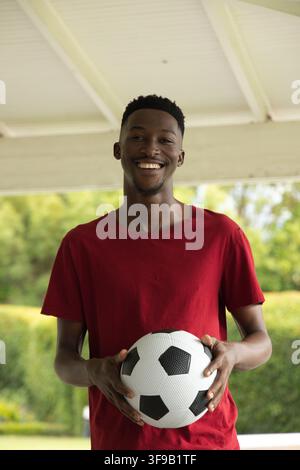 Uomo in piedi sotto un padiglione con tetto bianco che tiene un pallone da calcio bianco e nero all'altezza della vita, indossa una maglietta rossa Foto Stock