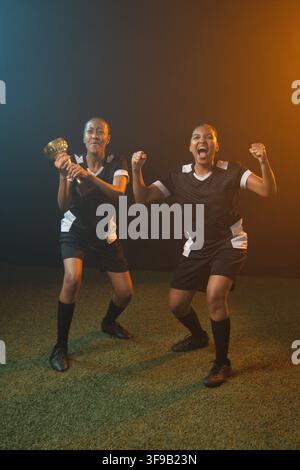 Donne afroamericane in tute nere che tengono un trofeo d'oro e applaudono sul campo erboso sotto luci blu-arancio Foto Stock