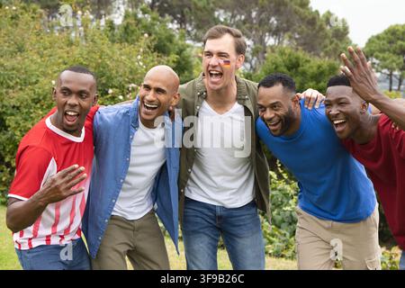 Diversi amici maschi che esultano e si sporgono verso la fotocamera nel parco indossando maglie e trucco facciale Foto Stock
