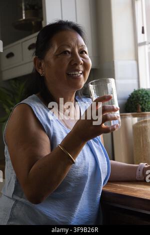 Donna asiatica anziana seduta a un bancone di legno vicino alla finestra con in mano un bicchiere trasparente e sorridendo Foto Stock