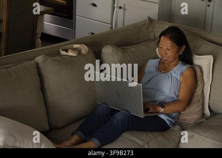 Donna anziana asiatica seduta su divano grigio a casa utilizzando un laptop argentato, cuffie beige su cuscino Foto Stock