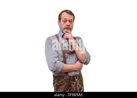 Un uomo anziano barbuto con indosso un lederhosen si trova in una posa pensierosa con la mano sul mento, isolato su uno sfondo chiaro. Foto Stock