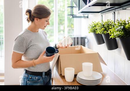 Donna prende tazze di ceramica da una scatola di cartone in un ambiente di cucina soleggiato Foto Stock