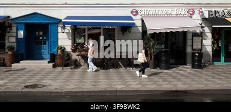 Due giovani donne passano davanti a un bar shawarma e a delle terrazze di caffè su Starotolmachevsky Lane a Zamoskvorechye, Mosca, Russia Foto Stock