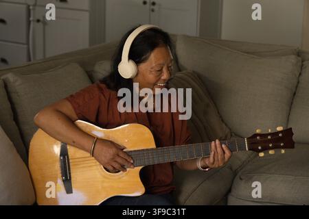 Donna anziana asiatica seduta su un divano grigio che suona la chitarra, indossa una maglia ruggine, auricolari bianchi Foto Stock