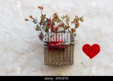 Cesto appeso in vimini con bacche e una forma a cuore rossa montata su uno sfondo murale ruvido Foto Stock