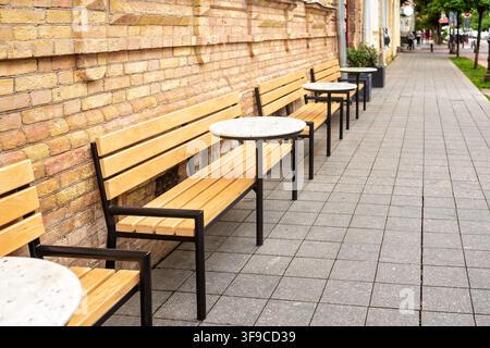 Terrazza all'aperto del caffè vuota con panche di legno e tavoli rotondi lungo un muro di mattoni sul marciapiede di una strada cittadina che si allontana in lontananza Foto Stock